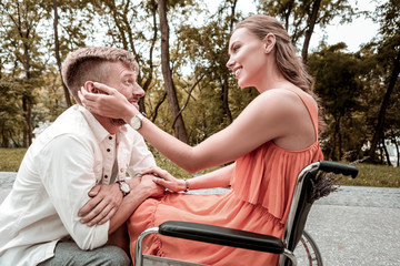 Being in love. Kind loving young girl sitting in the wheelchair in the park and smiling while...