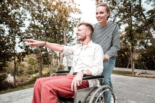 Happy man. Confident young man sitting in the wheelchair and pointing to the distance while showing beautiful park to his beloved woman