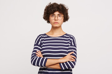 Closeup of calm serious emotionless young curly brownhair woman wears white and blue stripped sweatshirt and transparent black-framed round glasses keeps arms crossed isolated over white background - obrazy, fototapety, plakaty