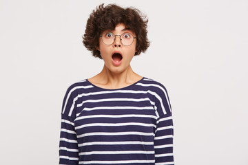 Studio shot of young beautiful girl looks amaized with open mouth, wears white and blue stripped sweatshirt and transparent black-framed round glasses isolated over white background