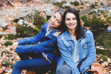 Two young girls friends sitting on a rock near the river in the autumn forest. Walk in the woods. Girlsfriends hiking at fall.