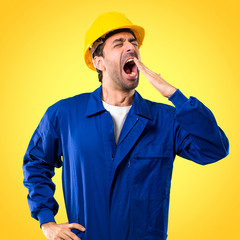 Young workman with helmet yawning and covering wide open mouth with hand. Sleepy expression on yellow background