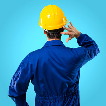 Young Workman With Helmet On Back Position Looking Back While Scratching Head On Blue Background