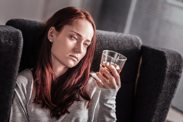Alcoholic drink. Cheerless unhappy young woman sitting at home and looking at the glass while...