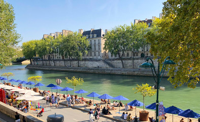 Paris, France, September 1, 2018: Locals gathered under blue umbrellas. Beach on Seine river in the center of Paris, - obrazy, fototapety, plakaty