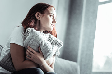 Thinking of you. Low-spirited girl holding pillows in both hands while sitting on the couch and...