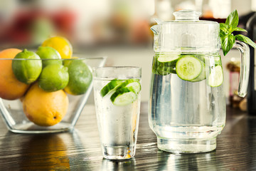 Pitcher with glass of cucumber water with bowl of fruits on the background.