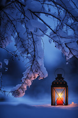 Candle lantern under the snowy branches at dusk. Christmas time in a wintery garden.
