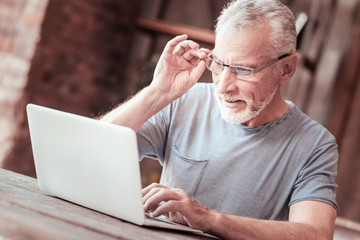 Obraz premium Full of interest. Close up of attentive elderly man touching his glasses rim while looking at the laptop screen and expressing concern