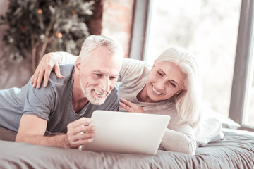 Having rest. Portrait of amused elderly family lying on the bed while hugging and using a laptop