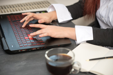 a woman's hand on a black-and-red laptop keyboard, which lies on a dark table, next to coffee and an open notebook