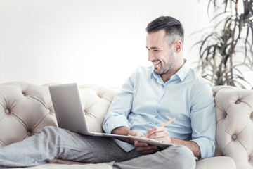 Making notes. Positive delighted brunette keeping smile on his face and turning head while staring at screen of his computer