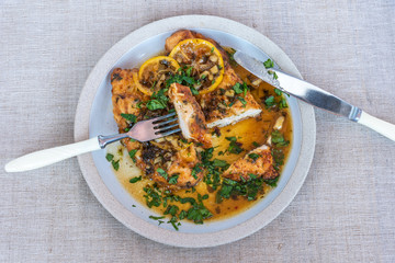 Chicken francese - Fried chicken breast with lemon slices and delicious sauce on a white plate on the table with a napkin, fork and knife, top view