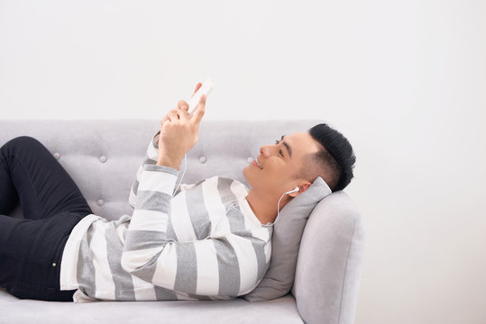Handsome guy in earphones is using a smart phone and smiling while lying on couch at home