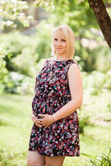 Fototapeta premium Beautiful pregnant woman in short floral maternity dress looking dreamy straight in the camera and smiling