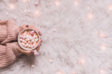 Cozy background. Hands holding mug of hot cocoa or hot chocolate with marshmallow on white rug.