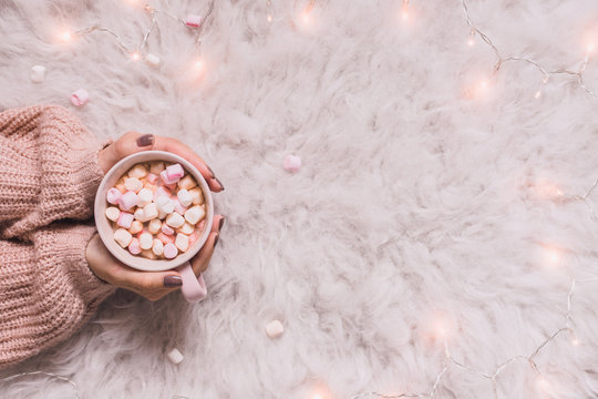 Cozy Background. Hands Holding Mug Of Hot Cocoa Or Hot Chocolate With Marshmallow On White Rug.