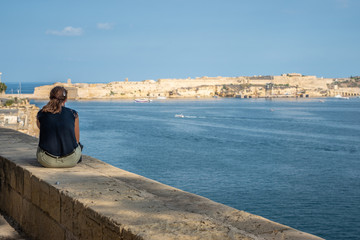 Fototapeta na wymiar Wide angle view of a young lady sitting on the harbour wall looking looking at the view. Horizontal. Malta Grand hourbour