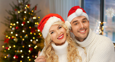 Fototapeta na wymiar holidays, celebration and people concept - smiling couple in sweaters and santa hats over living room with christmas tree background