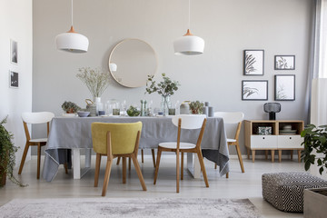Pouf, plants and round mirror in bright dining room interior with chairs at table under lamps. Real photo - obrazy, fototapety, plakaty