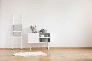 Real photo of white room interior for child with fluffy rug, ladder with coverlet and cupboard with elephant and boxes. Empty place for your crib