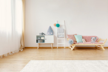 Scandi child room interior in real photo with carpet on the floor, window with drapes, cupboard with raindrop cushion and lounge with blanket and pompom