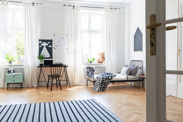 Striped carpet and windows in white teenager's room with black chair at desk near bed. Real photo