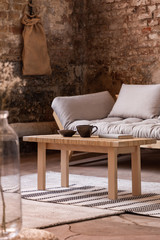 Wooden table on rug in front of grey settee in industrial interior with brick wall. Real photo