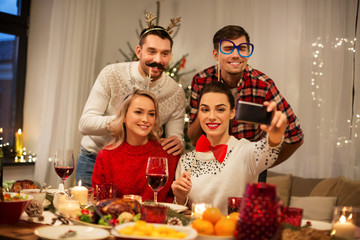 holidays and celebration concept - happy friends with party props taking selfie by smartphone at home christmas dinner