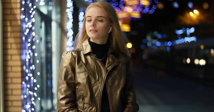 Attractive long-haired blonde young caucsian girl happily smiles straight to camera. City lights on the background. Positive mood, being happy, cheerfulness. Female portrait. wireless earphones