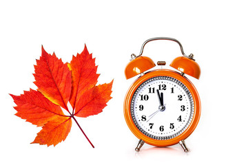 Orange alarm clock, watch hands showing midnight or noon, isolated on white background with autumn leaf. 