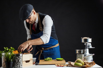 Food lover man adds lettuce on burger, cooking tasty burger with beef patty, vegetables and greenery over black background. Restaurant, Fast Food and Homemade food concept
