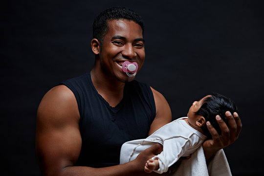 crazy afro father with a dummy in his mouth trying to calm down his baby. hysterical guy has beebn left along with his infant. daddy like a second chilf. childish behaviour. madness