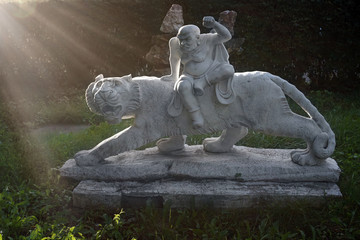 Marble sculpture of chinese tiger and warrior in a garden with green grass and sunset sun as background