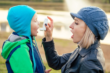 woman using an asthma inhaler to her son in a cold winter autumn Park