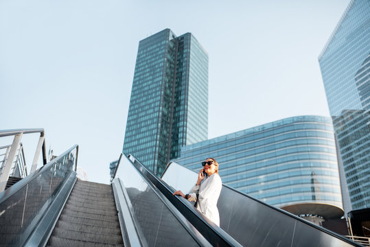 Stylish businesswoman in white suit talking phone while going up on the escalator at the business centre outdoors in Paris