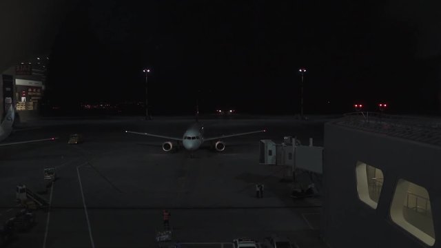 Aircraft is preparing for boarding, approaching to jet bridge in night. Engineers are walking around, servicing a plane, view from airport window