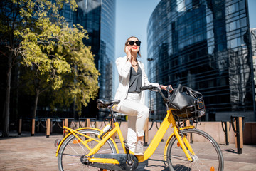 Portrait of a stylish business woman in white suit standing with bicycle at the financial district with modern buildings on the background