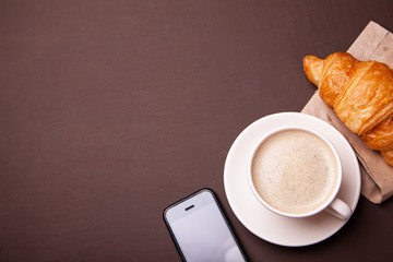 Coffee break, morning coffee. Cup of coffee with foam and croissant with smartphone.
