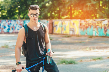 stylish man with bottle of water posing with bmx bike on summer park