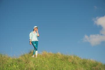 Naklejka na ściany i meble Fitness runner woman listening to music on the nature. Portrait of beautiful girl wearing earphones earbuds and running cap. against the background of a green field and blue sky