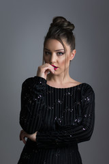 Fototapeta premium Beautiful elegant young woman in shiny black dress, posing, biting her fingers. Studio lighting, dark gray background.