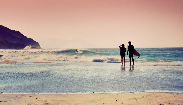 A man and a woman is standing on the beach looking at the ocean. Surfer is catching a wave in the background. Surfing concept. Surfing couple.