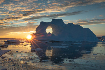 Sunburst behind a massive iceberg with a hole in it during the midnight sun season. Disko bay,...