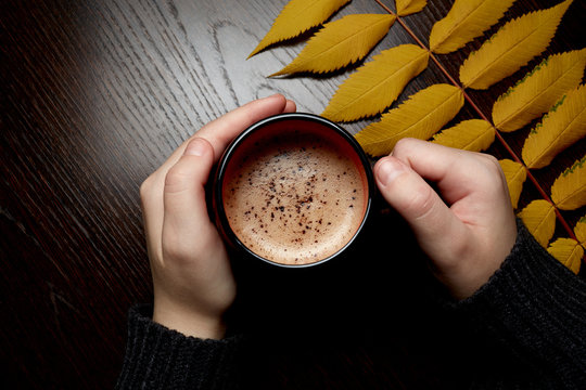 women's hands in a gray sweater holding a Cup of coffee with foam and chocolate on a brown wooden table on which lies a yellow autumn leaf
