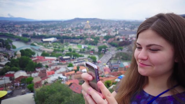 Young beautiful tourist girl is taking pictures of cityscape on smartphone camera and smiling
