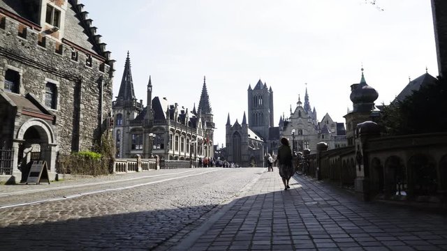 Young woman walking in historical centre of european city. Wide-angle view woman in silhouette go away on pavement road, medieval buildings at background St. Michael Bridge, Ghent