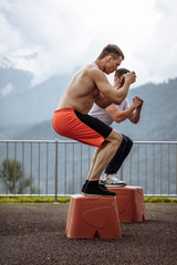 Fit young caucasian cross fit male instructor and his sportive student jumping on blocks at functional outdoor workout on fresh mountain air. Active and Health Life Concept