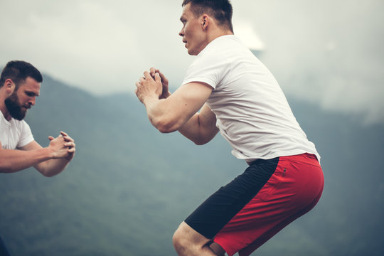 Jumping squats exercise - young handsome caucasian athletes doing functional outdoor workout in mountain area, training physical endurance and strength.