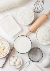 Fresh dairy products on white table background. Glass of milk, bowl of flour, sour cream and cottage cheese and eggs. Steel whisk and rolling pin. Top view